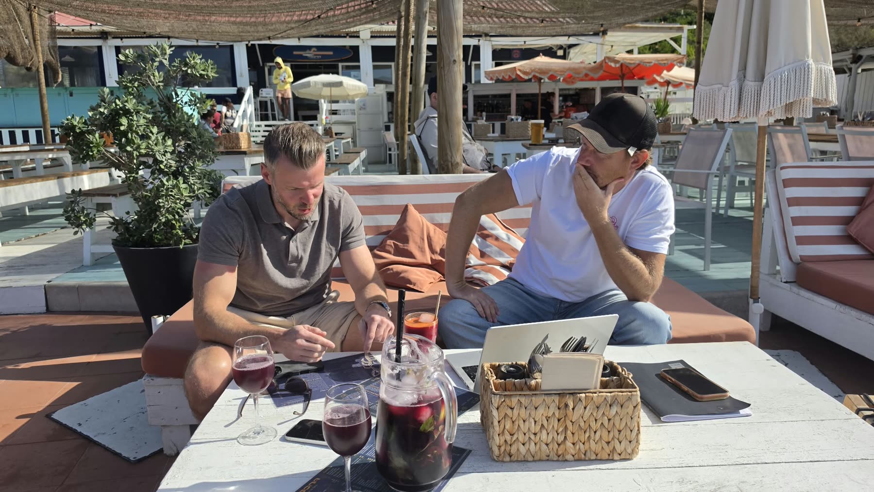Kris en een gast aan een lage tafel op een strandterras in Portugal, met een laptop, papieren en een kan sangria, diep in gesprek.